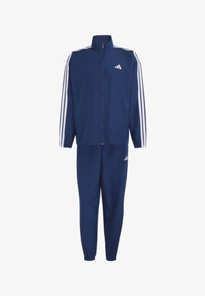 Junger Mann steht in einem dunkelblauen Adidas Trainingsanzug mit weißen Streifen und passenden Sneakers vor einem hellen einfarbigen Hintergrund.