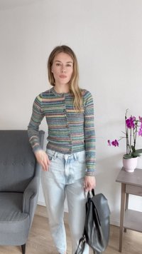 Cardigã riscado em cores pastel, com fecho frontal, combinado com jeans azul claro, segurando uma bolsa preta. Cadeira cinza e flores rosas ao fundo.