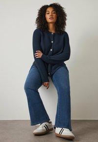 Cardigan de punto en color marino con seis botones redondos claros, combinado con Jeans de mezclilla oscuros acampanados y zapatillas blancas con rayas negras.
