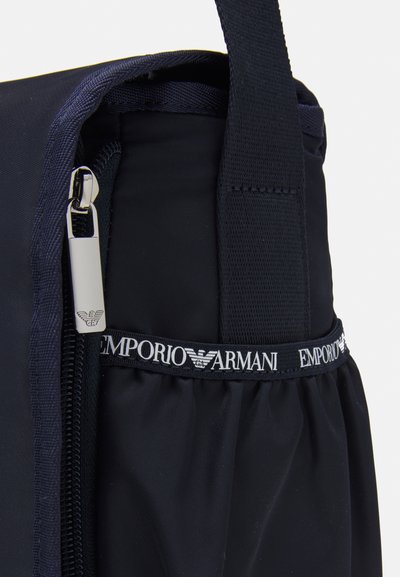 Tamsiai mėlyna nailono krepšys su užtrauktuku, tekstūruota rankena ir juodai baltu "Emporio Armani" logotipo apvadu aplink viršų.