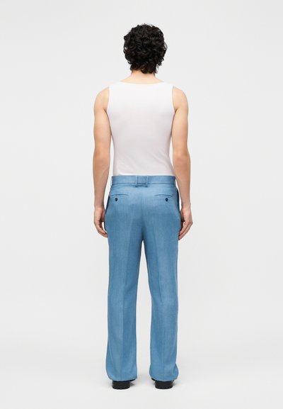 Homme aux cheveux bouclés portant un haut blanc sans manches et un pantalon bleu clair, debout de dos sur un fond uni.