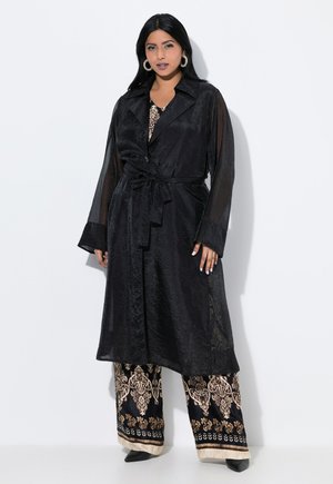 Cappotto lungo nero con cintura in vita e maniche trasparenti, abbinato a pantaloni a gamba larga con un motivo floreale dorato e bordi neri.