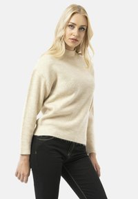 Crème kleurige ribbeltrui met een hoge halslijn, lange mouwen en een iets verkorte onderkant, gecombineerd met zwarte jeans en contrasterende stiksels.
