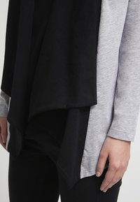 Schwarzes und graues, geschichtetes Oberteil mit geschwung hem. Weicher Stoff, lange Ärmel und lockere Passform. Mit einem drapierten schwarzen Überwurf.