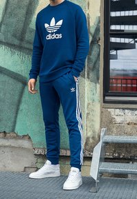 Sweat-shirt bleu avec le logo Adidas blanc, assorti à un pantalon de jogging bleu avec des rayures blanches sur les côtés. Des sneakers blanches complètent la tenue.