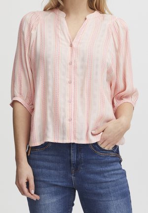 Femme portant un chemisier rose clair boutonné avec des rayures verticales et des manches trois-quarts, associé à un jean bleu avec des poches cloutées.