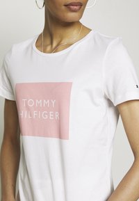 Camiseta de algodón blanca con un gráfico rectangular en color rosa claro que dice "TOMMY HILFIGER" en letras blancas y en negrita. Manga corta y escote redondo.
