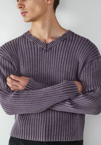 Junger Mann mit verschränkten Armen, der einen gerippten, lila V-Ausschnitt-Pullover trägt, vor einem schlichten hellen Hintergrund.