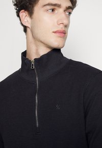 Zwarte ritsjumper gemaakt van textuurstof, met een hoge kraag en een klein logo op de borst. De rits voegt een functioneel detail toe.