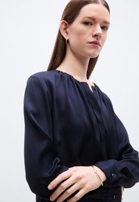Μπλούζα από μπλε σατέν σκούρο navy με μακριά μανίκια, κουμπιά, στρογγυλή λαιμόκοψη με ζαρντιέρες και λείο φινίρισμα, συνδυασμένη με ένα απλό ασημένιο βραχιόλι.