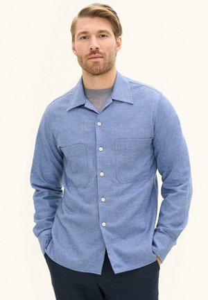 Mann mit hellbraunen Haaren und Bart, trägt ein geknöpftes hellblaues Hemd über einem grauen Oberteil, steht mit Händen in den Taschen vor einem schlichten Hintergrund.