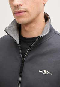 Chaqueta de cremallera de color gris oscuro con un cuello gris en contraste, que presenta un logo y el año "1962" en el pecho. Fabricada con un tejido suave.