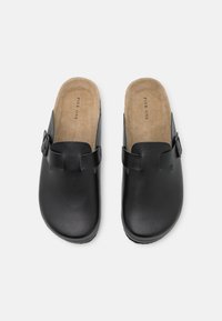 Sabots en cuir noir avec une finition lisse. Caractérisés par une boucle décorative et une semelle intérieure en daim beige. Design à bout rond et à arrière ouvert.