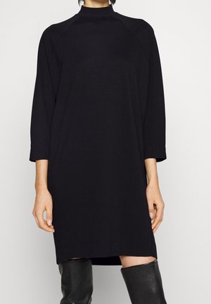 Kvinde iført en sort knælange kjole med lange ærmer kombineret med knæhøje sorte læderstøvler mod en neutral baggrund.