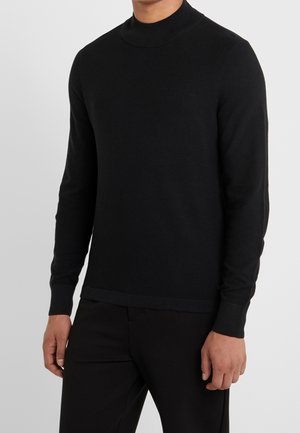 Mann trägt einen enganliegenden schwarzen Langarm-Pullover mit Stehkragen und eine schwarze Hose, steht vor einem schlichten hellen Hintergrund.