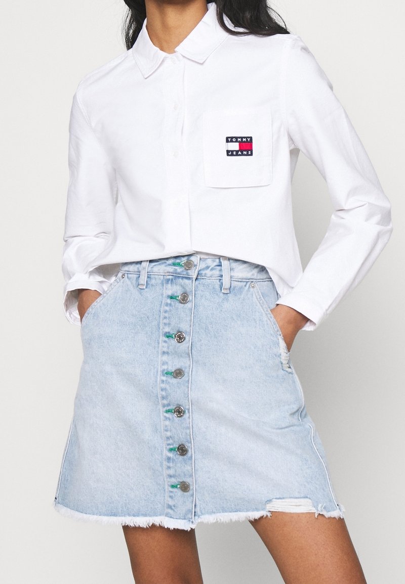 White long-sleeve button-up shirt with a chest pocket, paired with a light blue denim skirt featuring front buttons and a frayed hem.