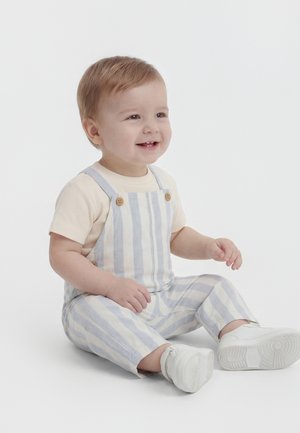 Małe dziecko o jasnych włosach, ubrane w kremową bluzkę, niebiesko-białe pasiasty ogrodniczki i białe trampki, siedzi i uśmiecha się na jednolitym tle.