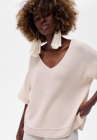 Vrouw met krullend haar in een oversized crèmekleurige gebreide top en grote witte kwastoorbellen, poseert met één hand in de zak tegen een witte achtergrond.