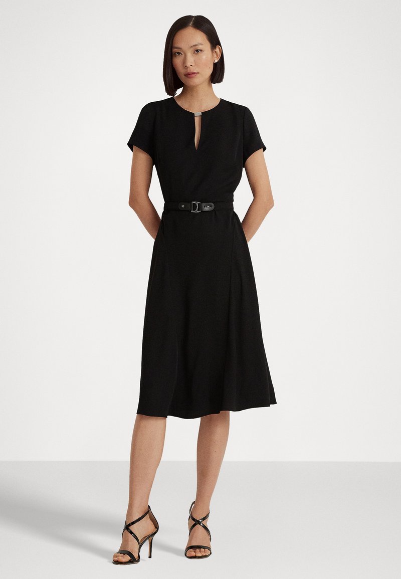 Abito nero con maniche corte, dettaglio del collo a chiave, vita aderente con cintura, gonna fluttuante. Modella in posa con tacchi a strappo neri.