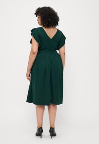 Robe verte foncée avec des manches courtes à volants, décolleté en V au dos, jupe pliss�ée et fermeture à glissière dissimulée. Portée avec des talons noirs. Texture de tissu lisse.