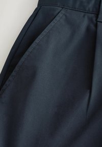 Nahaufnahme von dunkelblauen Stoffhosen mit schräger Seitentasche und sichtbaren Nahtdetails.