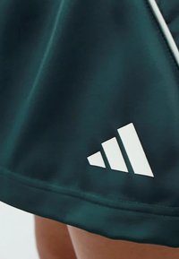 Tmavě zelená sportovní sukně s hladkým povrchem a bílým logem Adidas, se třemi pruhovanými akcenty podél spodního lemu.