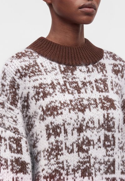 Personne portant un pull en maille blanc et marron avec un motif texturé et abstrait et un col côtelé marron uni.
