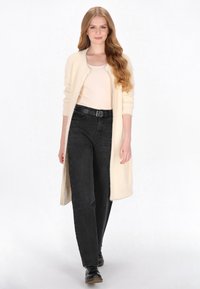 Cardigan lung crem, din tricot texturat, purtat peste un top bej și blugi negri cu talie înaltă, completat cu pantofi negri.