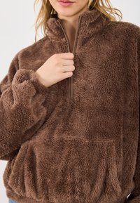 Brązowy polarowy bluza z wysokim kołnierzem, zamkiem błyskawicznym do połowy oraz kieszenią z przodu. Wykonana z miękkiego, teksturowanego materiału.