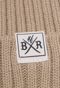 Beige strikket stof med et kvadratisk vævet label, der har bogstaverne "B", "R" og et grafisk element, tekstureret med ribbede mønstre.