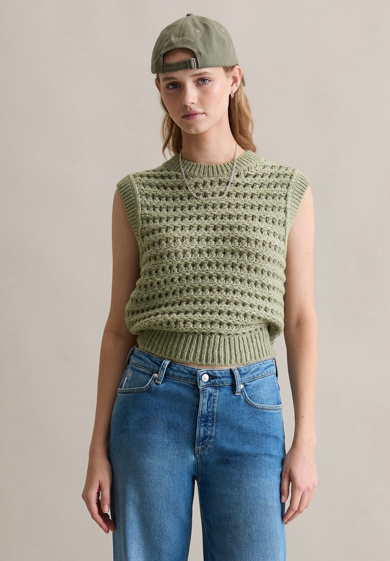 Groen gebreid sweater met korte mouwen en textuurpatroon, gecombineerd met lichtblauwe high-waisted jeans. Draag met een groene pet en zilveren ketting.