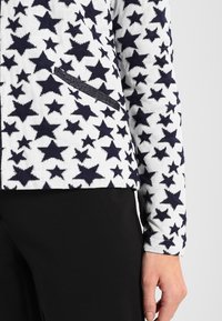 Fleecevest met marineblauwe sterpatronen op een witte achtergrond, voorzien van een zijzak en een gestructureerde stof.