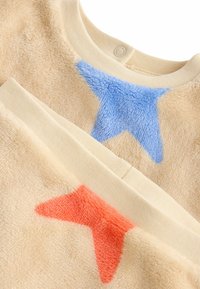 Beżowa bluza z polaru z aplikacjami w kształcie niebieskich i koralowych gwiazdek, okrągłym dekoltem oraz kontrastującym wykończeniem kołnierza. Miękka tekstura z puszystym wykończeniem.