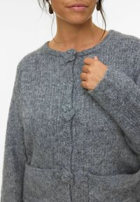 Cárdigan gris de punto con un acabado texturizado, que presenta un cuello redondo, un cierre central de botones y dos bolsillos frontales.