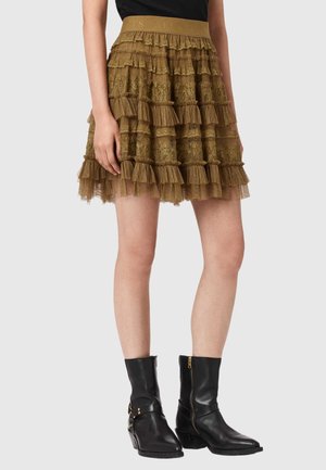 Kobieta ubrana w falbaniaste, oliwkowozielone mini spódniczki koronkowe i czarne skórzane botki za kostkę z zamkami błyskawicznymi i sprzączkami.