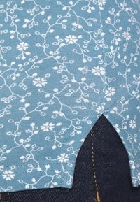 Blauwe stof met wit bloemrankpatroon, voorzien van een kleine zijsplit, gedragen over donker denim spijkerbroeken met zichtbare oranje stiksels.