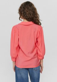 Blusa corallo con colletto, maniche a palloncino e una texture leggera. La vista posteriore mostra una vestibilità rilassata e uno stile casual. Abbinate a jeans blu.
