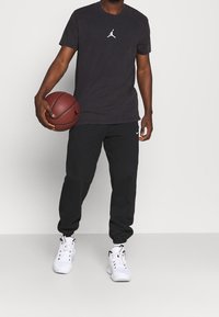 Man i svart Jordan-t-shirt och svarta Nike-joggingbyxor som håller i en basketboll, bärande vita träningsskor, stående mot en enkel bakgrund.