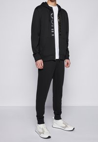 Felpa nera con zip davanti e tasche, t-shirt bianca sotto, pantaloni della tuta neri e scarpe da ginnastica bianche con dettagli grigi. Design elegante.