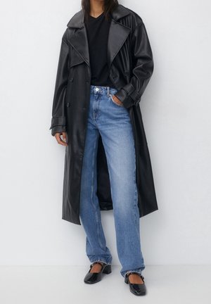 Personne portant un long trench-coat en cuir noir, un top noir, un jean bleu et des chaussures noires à bouts carrés et talons bas sur fond blanc uni.