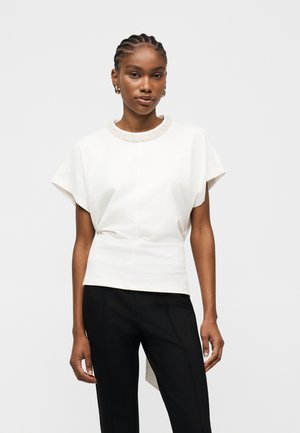 Donna con capelli intrecciati che indossa una maglietta bianca a maniche corte con scollatura decorata e pantaloni aderenti neri, in piedi davanti a uno sfondo semplice.