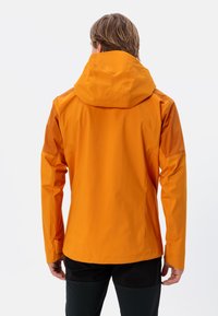 Oranje waterdichte jas met een capuchon, beschikt over naadloze schouders, verstelbare manchetten en een gladde afwerking. Draagt met zwarte broeken.