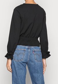 Top nero a maniche lunghe con vita elastica arricciata e polsini con volant, abbinato a jeans in denim blu a vita alta con tasche posteriori caratteristiche.