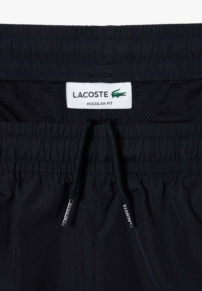 Shorts de bain noirs avec une taille élastique, fermeture à cordon, doublure en mesh et une étiquette Lacoste arborant un logo de crocodile vert.