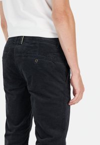 Zwarte corduroy broek met een textuurafwerking, voorzien van een achterzak en een knoopsluiting, zijkant getoond tegen een effen achtergrond.