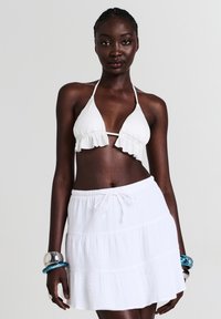 Bikini bianco con bordi arricciati e design a collo halter, abbinato a una gonna bianca a strati. La modella indossa bracciali argento e blu.