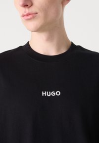 Melnas kokvilnas t-krekls ar apaļu kakla izgriezumu, ar balto izšūto 'HUGO' logo. Gluda tekstūra, īsas piedurknes un brīva piegriezuma. 