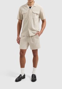 Lichtbeige shirt met korte mouwen en twee voorkruiden, gecombineerd met bijpassende korte broek. Ggedragen met zwarte loafers en witte sokken. Gestructureerde stof.