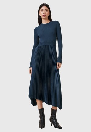 Langsleeve donkergroen jurk met een gestructureerde ribben bovenkant en een geplooide rok. Heeft een asymmetrische zoom en wordt gedragen met zwarte enkellaarsjes.