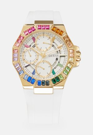Montre Guess ton or avec bracelet blanc, pierres précieuses colorées autour de la lunette et trois sous-cadrans sur un cadran blanc avec aiguilles dorées.